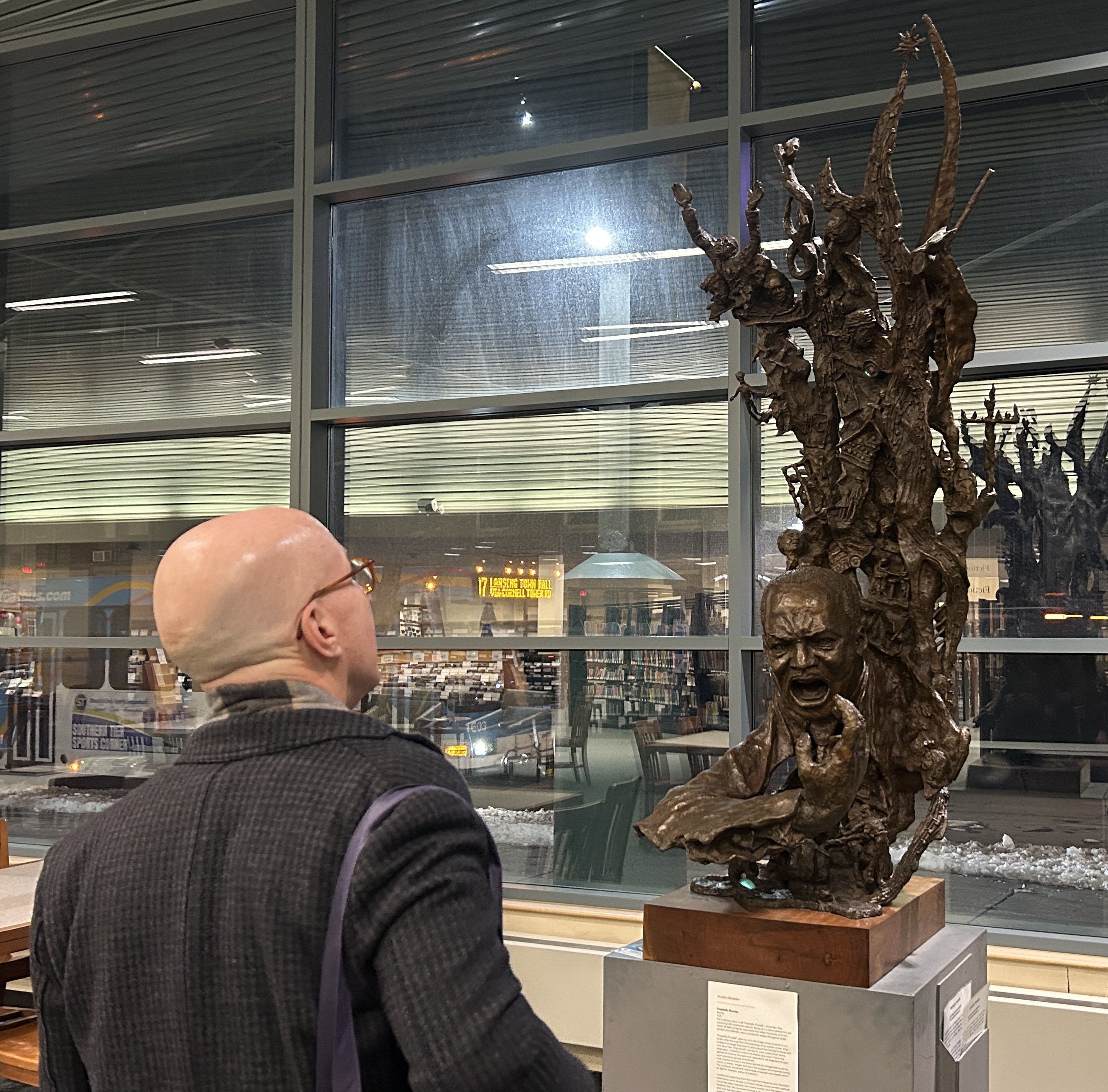 Author Jonathan Eig, wearing a gray overcoat and scarf, admires the sculpture Prophetic Thunder at TCPL. The sculpture shows Dr. Martin Luther King Jr, with his hand outstretched, delivering a fiery sermon. Above him is a representation of flame, with objects and people related to Dr. King's Life and the Civil Rights movement