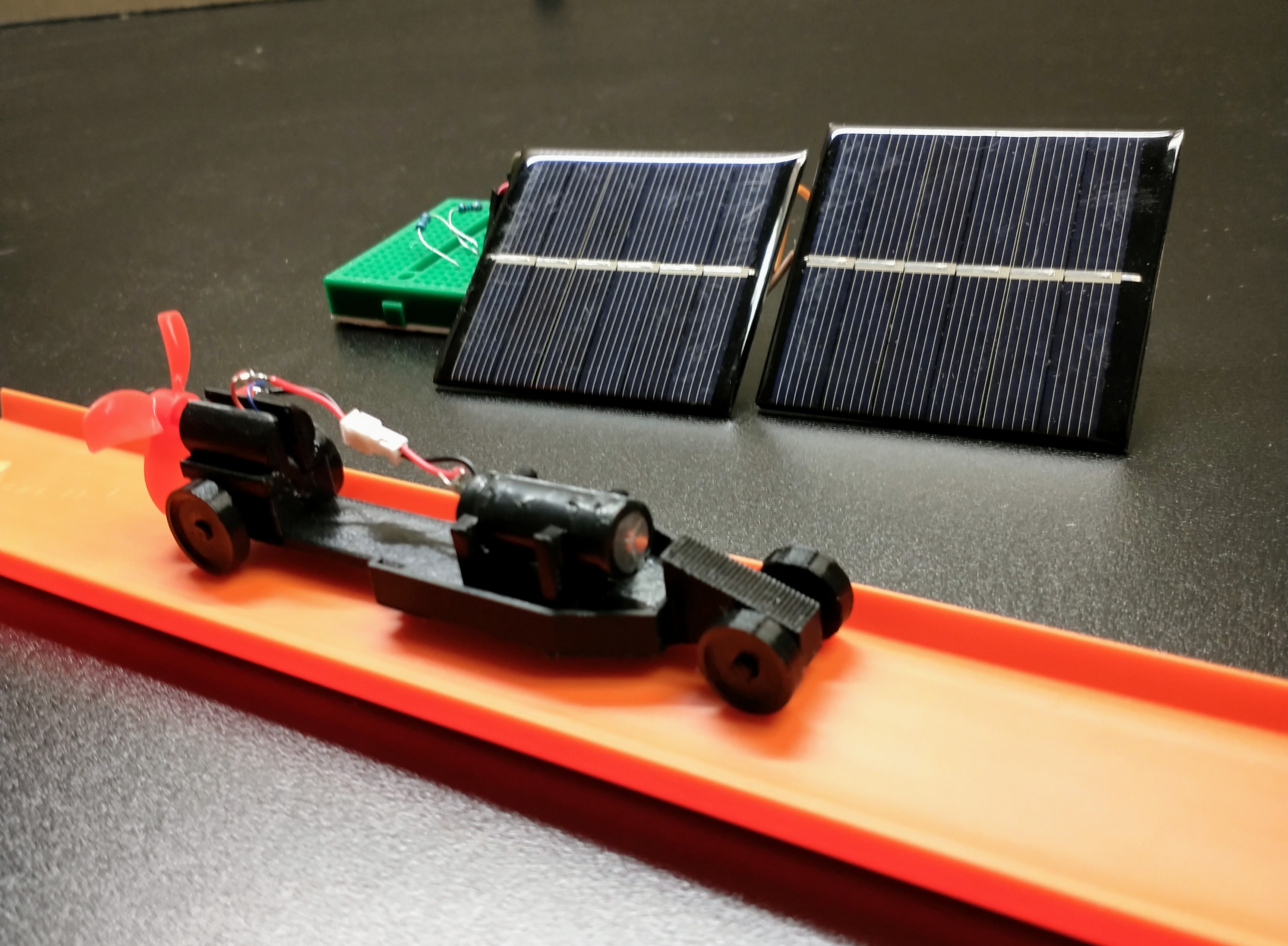 A 3D printed car on orange Hot Wheels track next to two solar cells.