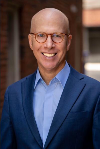 A photo of author Jonathan Eig. He is wearing small round glasses, a light blue Oxford style shirt and a dark blue suit jacket. Photo credit: Doug McGoldrick