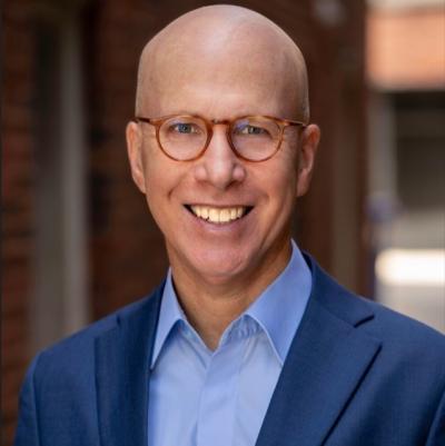 A headshot of Jonathan Eig, taken by photographer Doug McGoldrick. Eig is wearing a dark blue sport coat, a light blue oxford style shirt and small round horned-rim glasses 